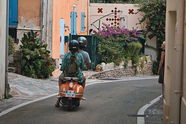 Two people riding a moped down an old, narrow street in Bormes-les-Mimosas, Provence, France
